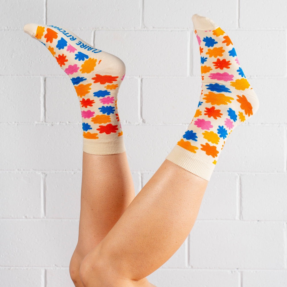A pair of cotton-blend socks designed by Claire Ritchie. A model wears a cream sock, dotted with flowers in turmeric, orange, pink and blue. The words Claire Ritchie are printed on the sole of the foot in blue. 