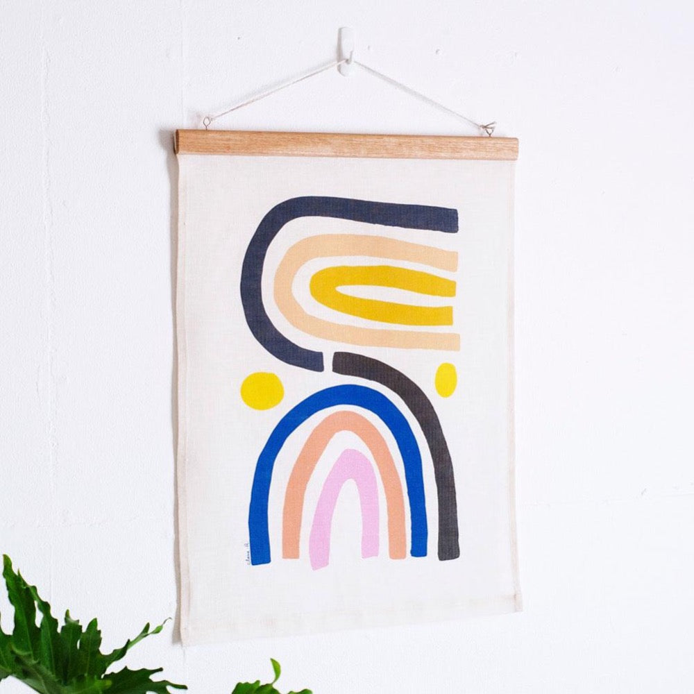 100% Linen Wall Hanging with original design by Claire Ritchie. Two multi-colour rainbows sit side by side with a yellow orb by each side. The hanging sits on a white wall with a green house plant in the bottom left of the image. 